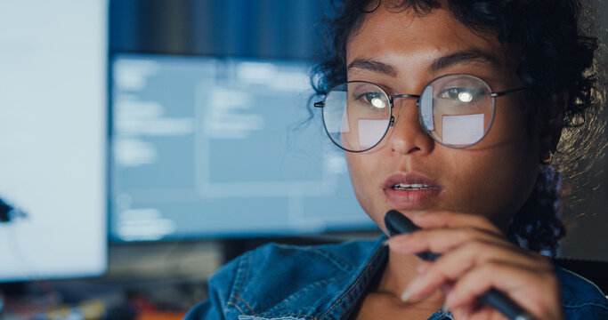 Close-up young Black female programmer coding AI software on multiple screens in night office. Network security, cybersecurity, machine learning, working late, overtime, robotics automation concept.