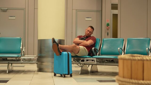 Air travel. A tired man waits for his delayed flight at the airport. The man rests his feet on his suitcase and dozes off while waiting.