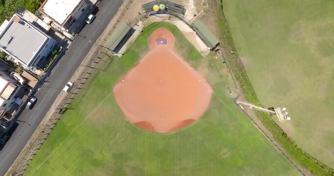 Top down aerial drone view of a single baseball field, showcasing the green grass outfield, red clay infield diamond, dugouts, and a very prominent blue curved outfield fence line. Zenithal shot.