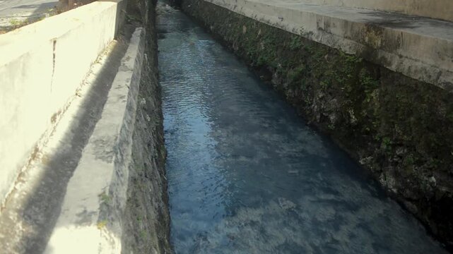 Narrow Concrete Water Canal With Flowing Dark Water And Green Mossy Sides In A Sunny Outdoor Setting