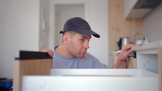 Professional handyman with toolbox performing maintenance in kitchen of home for repair project. Man wearing cap uses screwdriver to assemble wooden furniture cabinets in modern domestic interior.