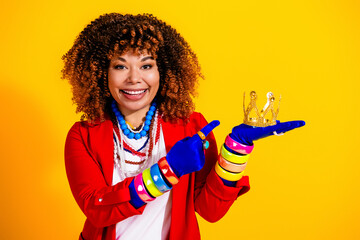 Happy woman in colorful fashion and accessories holding a small gold crown and smiling against a...