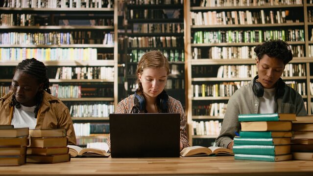 Young woman student with headphones around her neck taking notes on laptop at wooden study desk near blurred shelves, digital learning solo study in library workspace for academic learning themes.