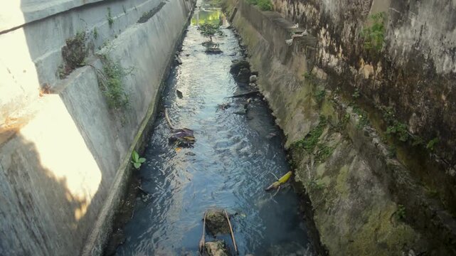 Narrow Urban Drainage Channel With Murky Flowing Water And Weathered Concrete Walls In City Alley