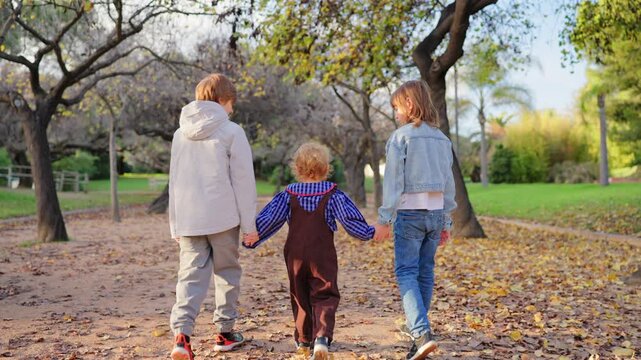 Three siblings walk together holding hands along a leaf-covered path in an autumn park. Older boy and girl guide a toddler in overalls through golden fall scenery with trees.