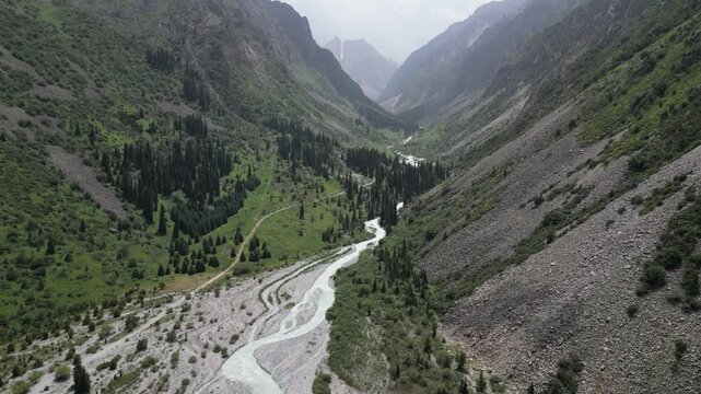 Aerial view of a winding river flowing through a deep mountain valley with steep rocky slopes, pine forests, and a dirt path in Bishkek, Bishkek City, Kyrgyzstan.