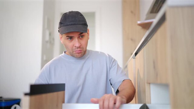 Maintenance and toolbox with skilled worker in kitchen of home for repair project. Man wearing cap uses screwdriver to fix cabinet drawer in modern interior with wooden cabinetry and bright lighting.