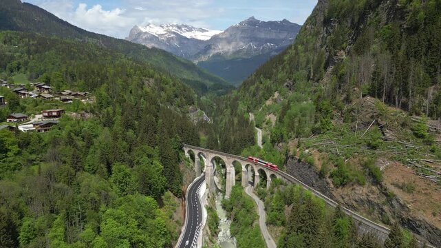 Aerial view of a red train crossing a historic stone viaduct surrounded by lush green forests and snow-capped mountains in Chamonix, Auvergne-Rhone-Alpes, France.