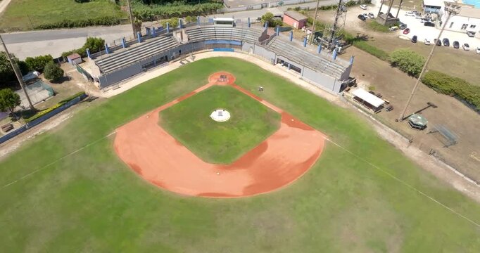 Aerial drone view of an empty baseball stadium on a sunny day, showcasing the distinctive red clay diamond, green grass outfield, concrete grandstands, pitcher's mound, and clear baseline markings.