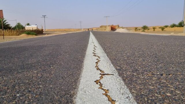 Long desert road in the Sahara near Timimoun, Algeria, marked by pronounced longitudinal cracks along the centerline, while pedestrians cross the highway on the distant horizon