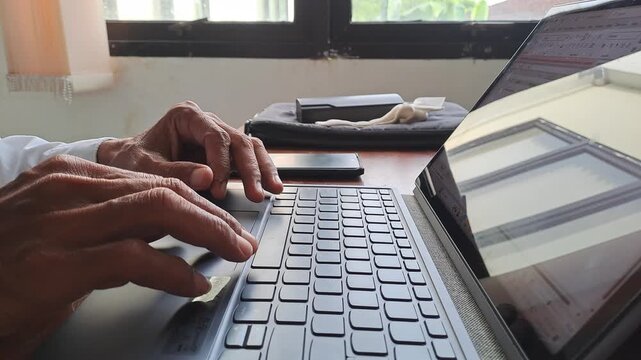 PAMEKASAN, INDONESIA - JANUARY 20, 2026: Close-up of the experienced hands of a mature Asian businessman or senior office worker diligently typing on a laptop keyboard. He is focused completing task.