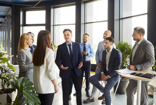 Mixed group of business people standing around male business leader and listening to him during corporate meeting or training lecture. Entrepreneur, brainstorming and teamwork with employees.