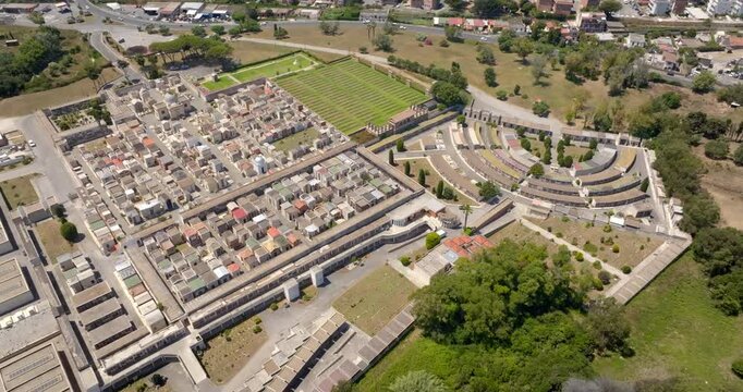 Aerial drone view of a large organized cemetery complex with dense rows of tombs, graves, and monuments next to structured green lawns and surrounding roads in a sunny summer morning.