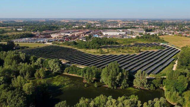 Aerial view of solar farm with rows of photovoltaic panels near a pond and industrial buildings in Saint-Denis-de-l'Hotel, Centre-Val de Loire, France.