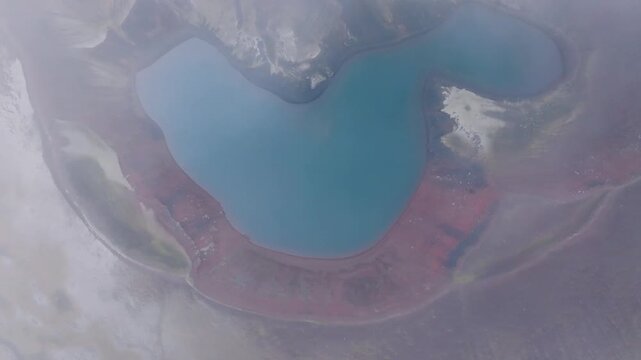 Aerial view of a volcanic crater lake with blue water and reddish mineral deposits surrounded by mist in Iceland.
