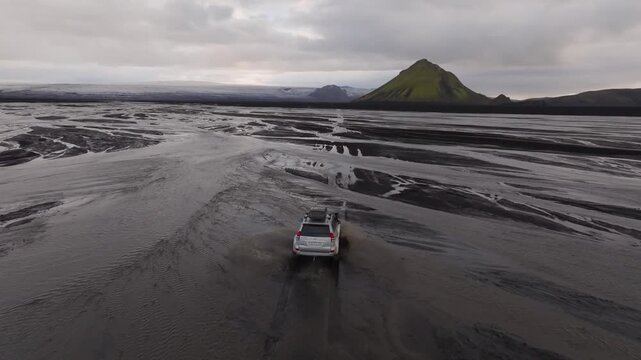 Aerial view of a 4x4 vehicle driving through a shallow river in the Icelandic highlands with a green mountain in the background under an overcast sky, Iceland.