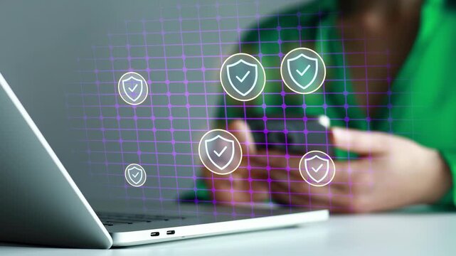 Person uses phone while sitting near a laptop with digital shields illustrating cybersecurity concepts.