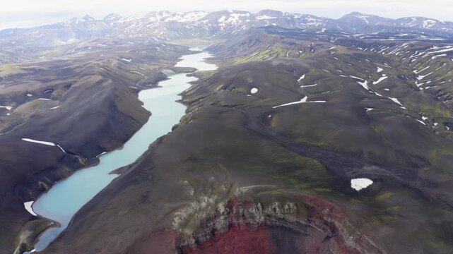 Aerial view of a turquoise glacial river winding through rugged volcanic mountains with patches of snow in Iceland.
