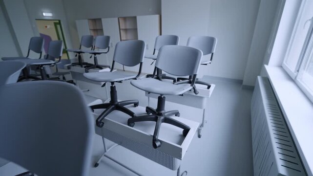 Chairs stand on desks in empty classroom near wide window. Clean minimalist interior suggests end of school day and room preparation