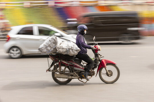 Hanoi, Vietnam - 11 November 2025: View of a motorcyclist carrying large heavy bags on a red motorbike navigating through busy city traffic with motion blur.
