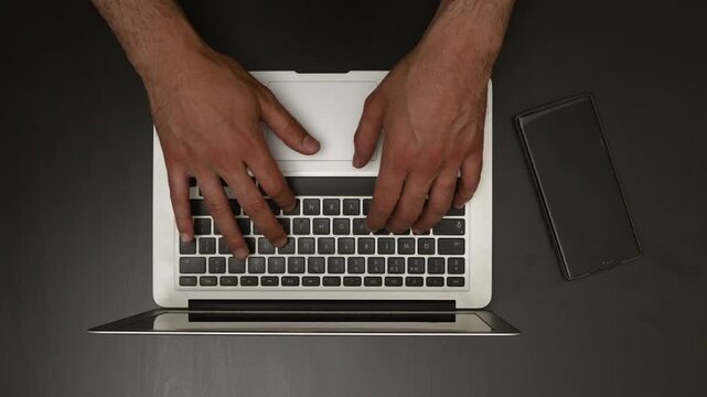 Top View of Man Typing on Laptop Keyboard at Dark Desk with Smartphone