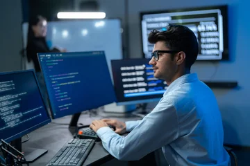 Young South Asian man in glasses works at a multi-monitor setup with code and data screens as a female colleague stands in the background. © Jestercine
