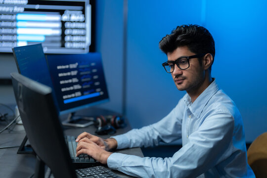 South Asian man in glasses types intently at his keyboard with a data dashboard glowing behind him against a deep blue office wall at night.