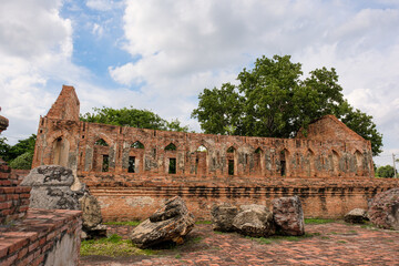 Fototapeta premium Wide exterior view of the ancient red brick assembly hall ruins at Wat Kudi Dao in Ayutthaya, Thailand. Large broken stone debris rests on a paved courtyard in front of arched windows and green trees