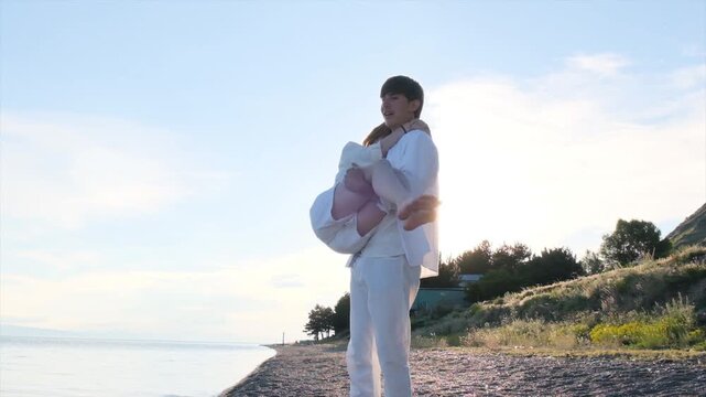Teenage boy carrying a girl in his arms on a seashore at sunset. Young couple, first love and sibling relationship concept as happy teenagers enjoy time together by the sea in white clothes.