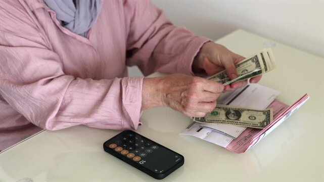 Senior woman counting cash at home with bills and smartphone calculator on the table. Close-up of retirement finances, household expenses and personal budget planning.