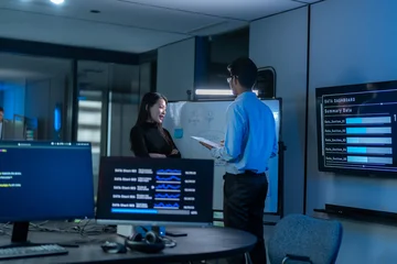 South Asian man presents at a whiteboard beside a wall screen showing a data dashboard while a woman listens in a dark blue office meeting room. © Jestercine