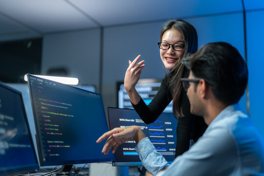 Asian woman in glasses gestures and laughs with a seated male colleague during a relaxed coding session, with code and data charts behind them.