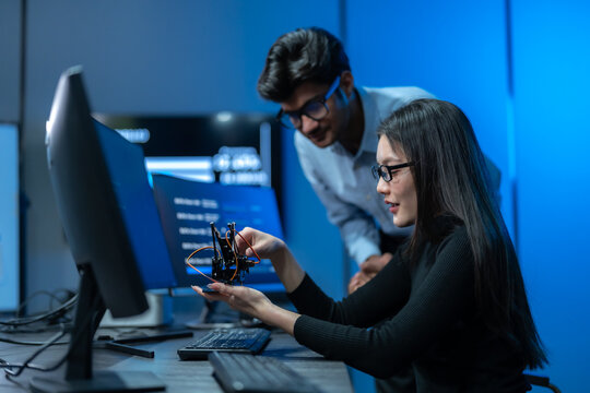 Asian woman holds a small robotic device while a male colleague leans in to inspect it together at a workstation in a dark blue robotics office.