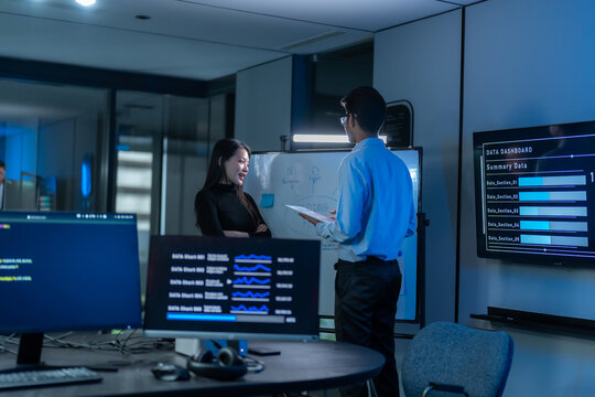 South Asian man presents at a whiteboard beside a wall screen showing a data dashboard while a woman listens in a dark blue office meeting room.