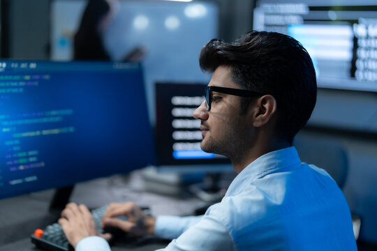 South Asian man in glasses studies code on his monitor while a colleague works nearby in a dark, blue-lit software development office.