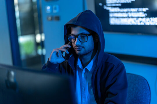 South Asian man in a hood and glasses talks on a phone while code glows behind him in a dark blue office, hinting at hacking and cyber fraud.