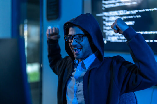 South Asian man in a hood and glasses raises both fists and shouts in triumph in front of code, suggesting a hacking breakthrough or win.