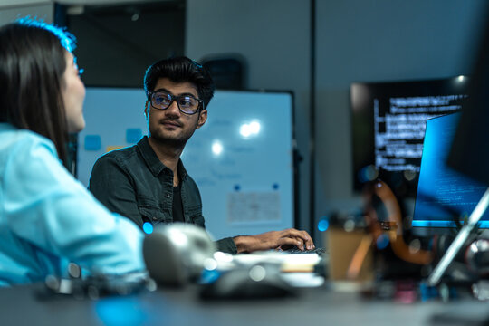 South Asian man in glasses types and talks with a colleague in the foreground at a desk with a whiteboard and lab equipment in a blue-lit office.