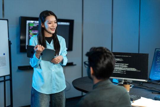 Asian woman in a blue shirt smiles and holds a tablet as she talks to a seated male colleague, with a whiteboard and code screens behind them.