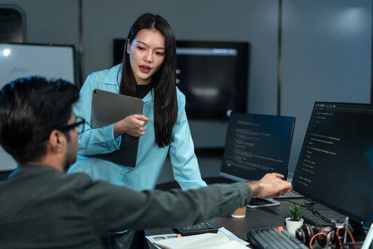 Asian woman in a blue shirt holds a tablet and discusses code with a seated male colleague who points at a monitor in a dark blue tech office.