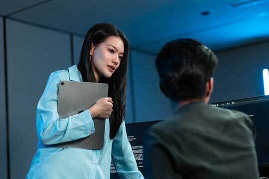 Asian woman in a blue shirt holds a tablet to her chest as she talks seriously with a male colleague seen from behind in a dark blue office.