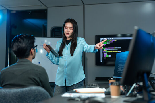 Asian woman in a blue shirt points toward a screen of code as she explains it to a seated male colleague in a dark blue tech office at night.