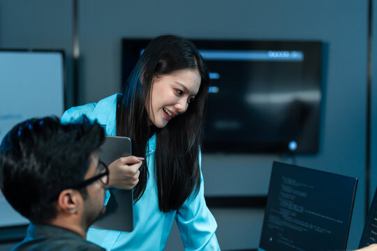 Asian woman in a blue shirt holds a tablet and smiles warmly as she leans toward a seated male colleague in a dark blue tech office at night.