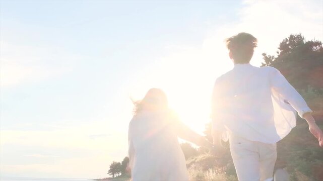 Camera following a carefree and happy teenage boy and girl running hand in hand toward the sunset along the seashore. First love, sibling relationship and joyful youth concept by the sea.