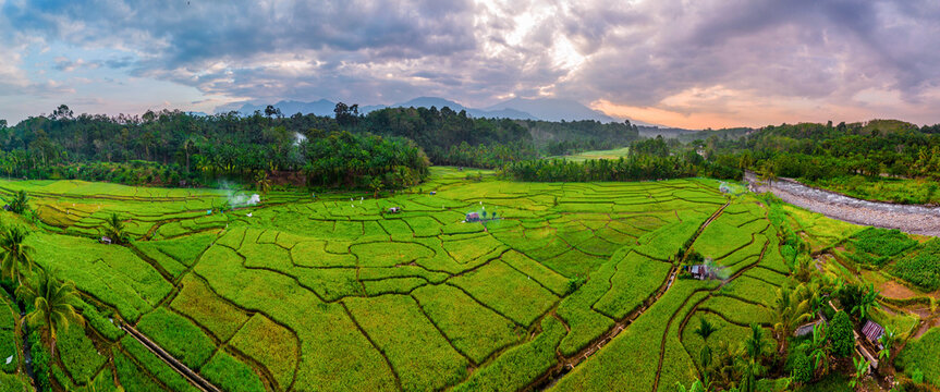 Aerial view of Taba Tembilang rice paddies with lush green terraces, a winding river, and distant mountains under a dramatic cloudy sky in Arga Makmur, Bengkulu, Indonesia.