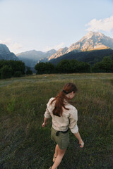 Obraz premium Woman lifestyle on a meadow with distant mountains, back view, dressed in casual shirt and shorts, exploring nature and enjoying peaceful outdoor scenery during golden hour.