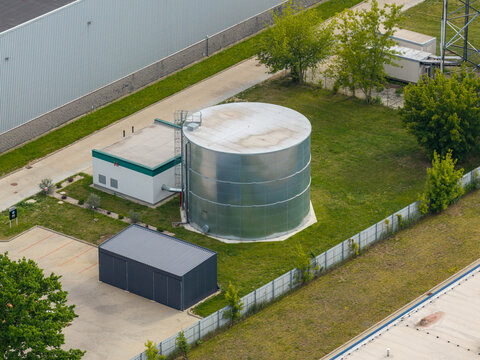 Aerial view of a large cylindrical metal water tank for fire safety systems situated in an industrial area with green grass and nearby buildings in Warsaw, Mazowieckie, Poland.