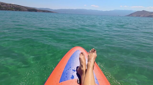 POV video of legs stretched out on a stand up paddle board drifting across emerald and turquoise Adriatic Sea water near Ba&scaron;ka on Krk Island, Croatia