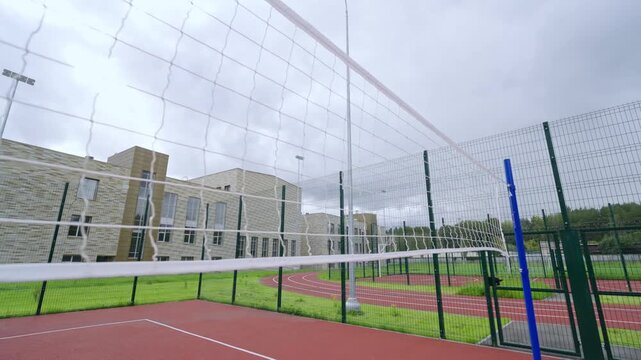 Volleyball net stretches across foreground of outdoor court. Modern school building stands behind green fence under cloudy autumn sky