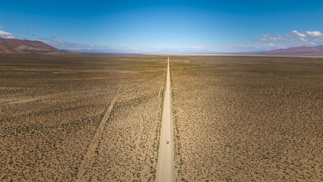 Aerial view of a long straight road cutting through the vast arid desert landscape under a clear blue sky with a single vehicle driving along the path, Argentina.